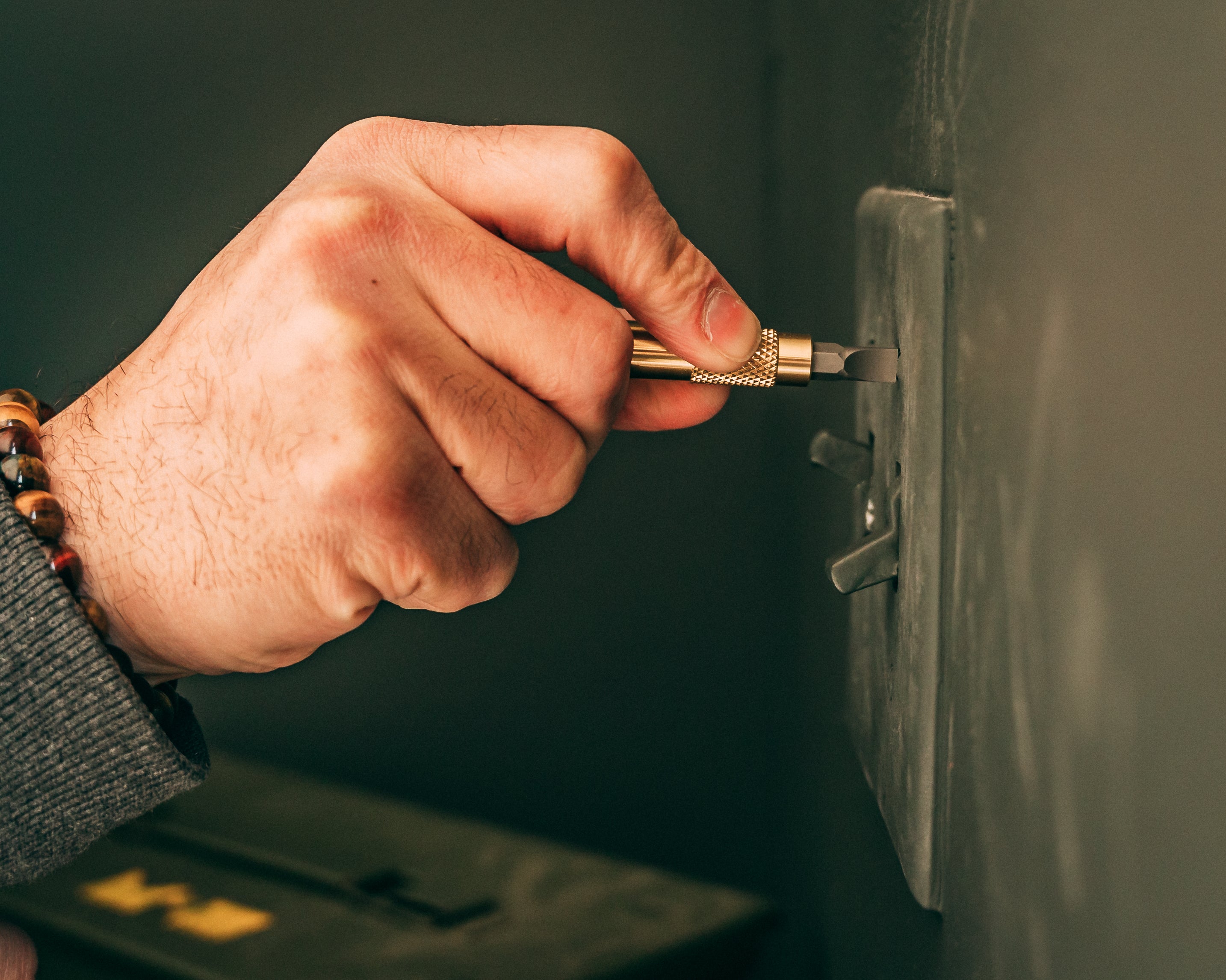 Hand holding brass screwdriver keychain with flat bit being used to turn screw in lightswitch cover.